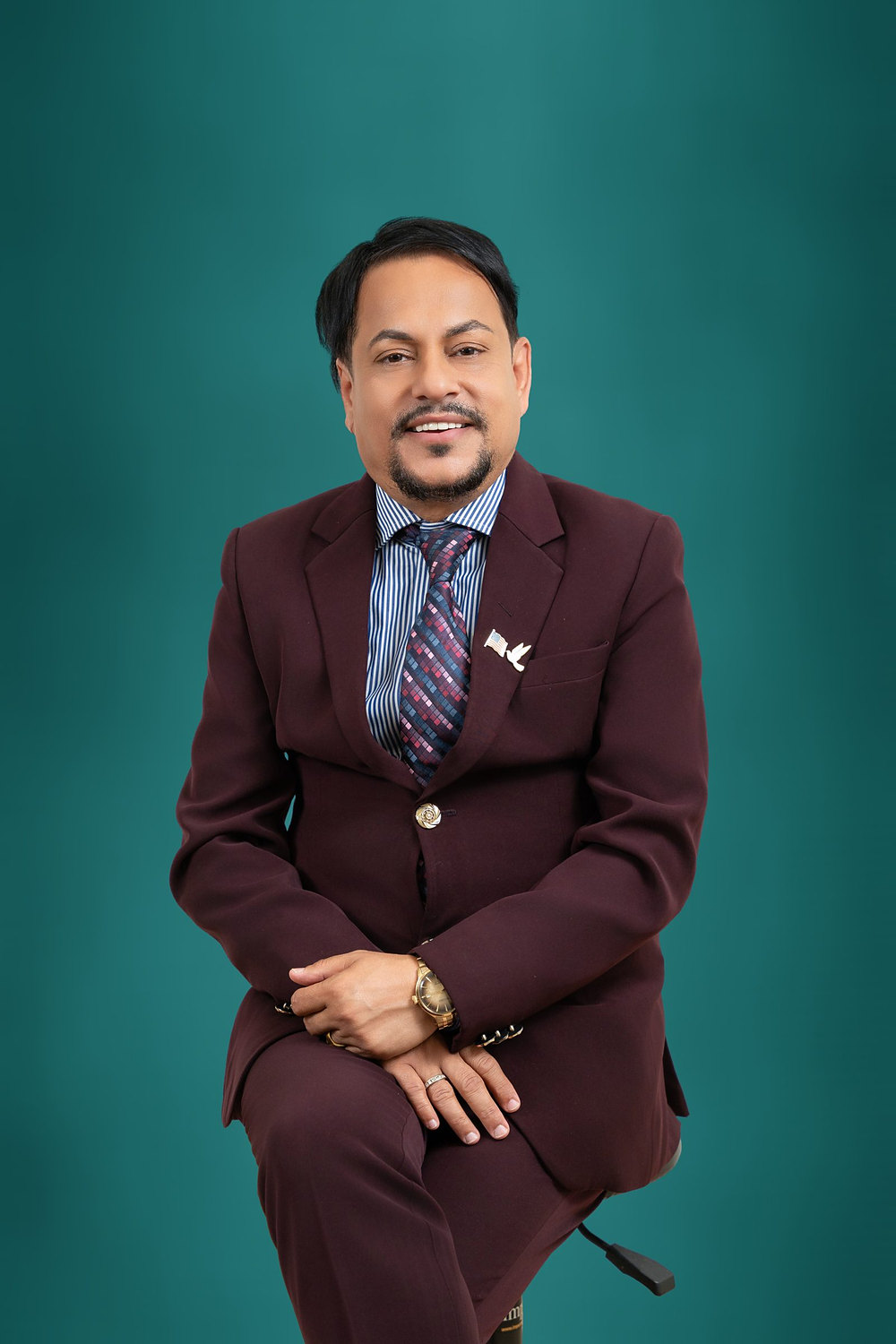 Studio headshot of a businessman in a burgundy suit and patterned tie against a teal backdrop
