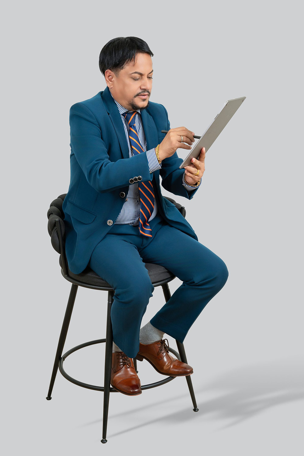 Studio portrait of a professional in a blue suit seated on a stool using a tablet, gray backdrop